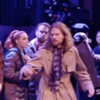 a man in a trench coat and plaid scarf stands in front of a group of people on a stage ..