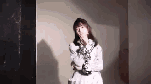 a woman in a white dress is standing in a room holding her hand to her chin .