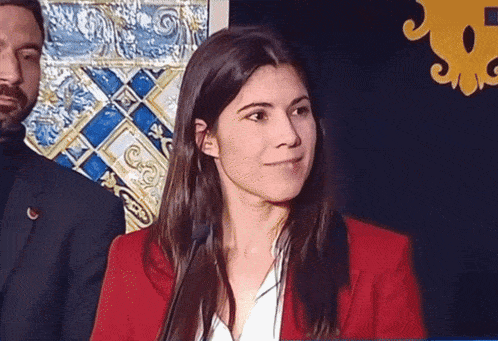 a woman in a red jacket stands in front of a microphone with a man in a suit behind her