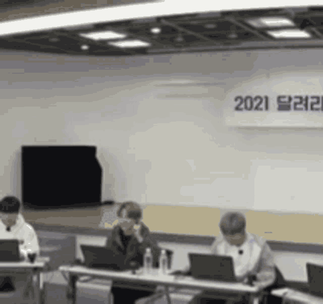three people are sitting at tables with laptops in front of a sign that says 2021 .