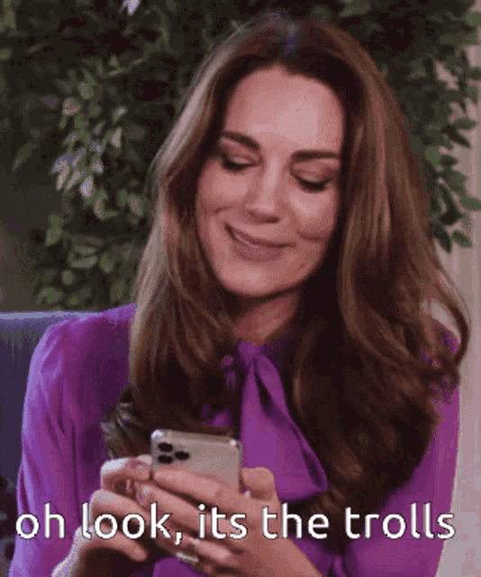 a woman in a purple shirt is smiling while looking at her phone with the words oh look it 's the trolls