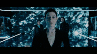 a woman in a black suit is standing in a dark room with two men behind her