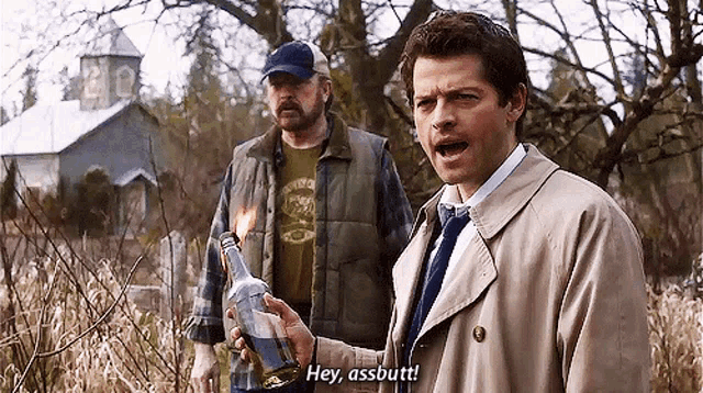 two men are standing in a field one is holding a bottle that is on fire and the other is saying hey assbutt