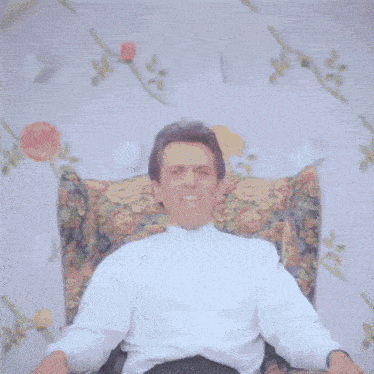 a man in a white shirt is sitting in a floral chair making a face .