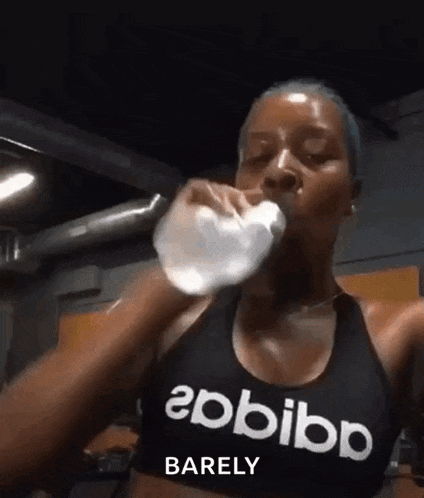 a woman is drinking water from a bottle in a gym while wearing a black adidas tank top .