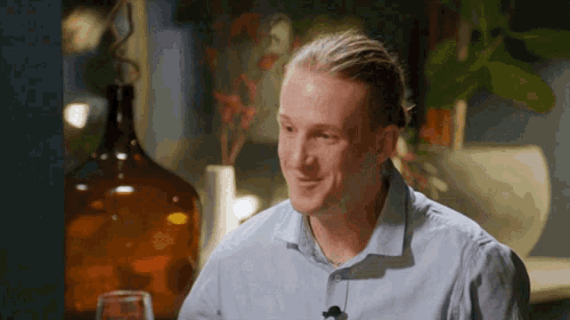 a man in a blue shirt is sitting at a table with a vase of flowers behind him .