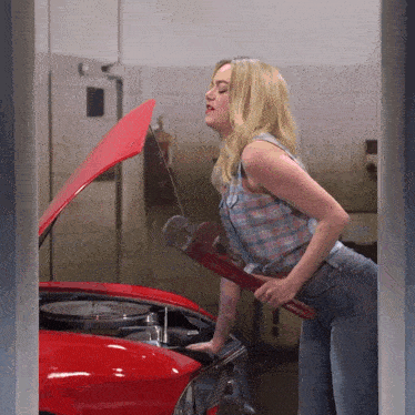 a woman in a plaid shirt is holding a wrench and looking under the hood of a red car