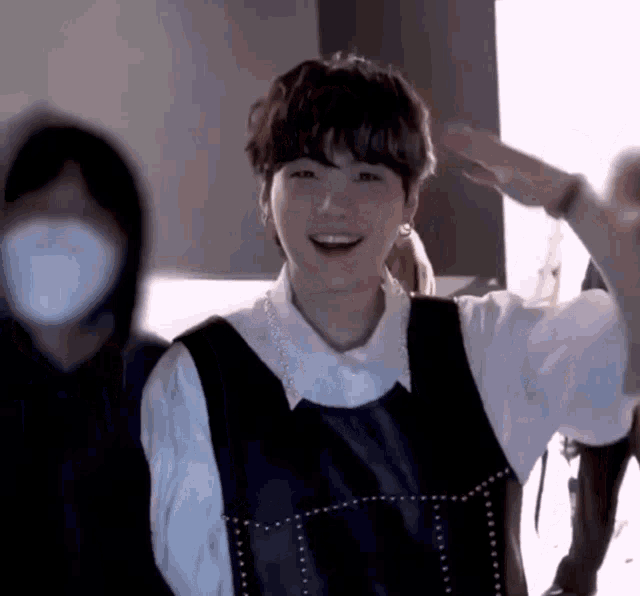 a young man wearing a white shirt and a black vest is smiling and waving his hand .