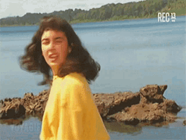 a woman in a yellow shirt is standing on a rocky beach near a lake .