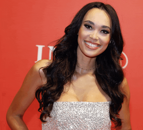 a woman in a strapless dress smiles in front of a red background with the letter l on it