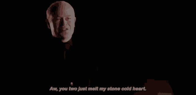 a man in a black suit and tie is talking in a dark room with his hands folded .