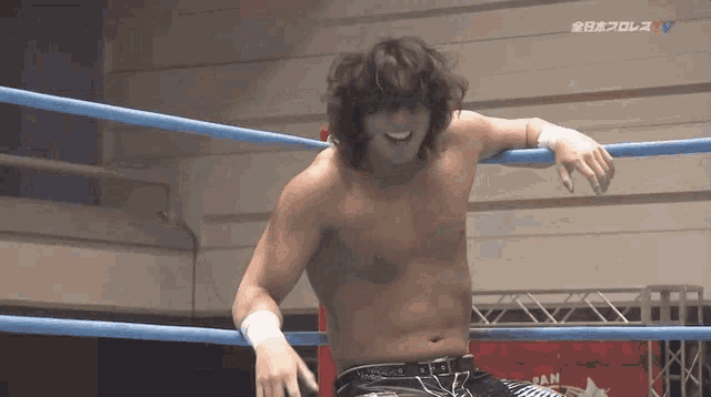 a shirtless wrestler leans on a blue rope in a wrestling ring .