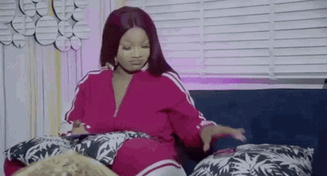a woman in a pink jacket is sitting on a couch with pillows and a cell phone .