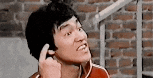 bruce lee is making a funny face while holding his finger to his ear in front of a brick wall .