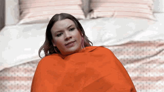 a woman wrapped in an orange blanket is sitting on a bed looking at the camera