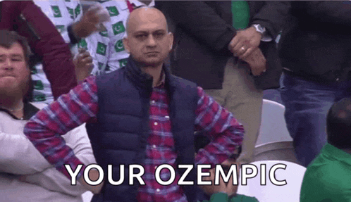 a bald man in a plaid shirt is sitting in a crowd with his hands on his hips and the caption your ozempic