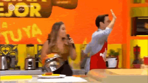 a woman is singing into a microphone while a man is dancing in a kitchen