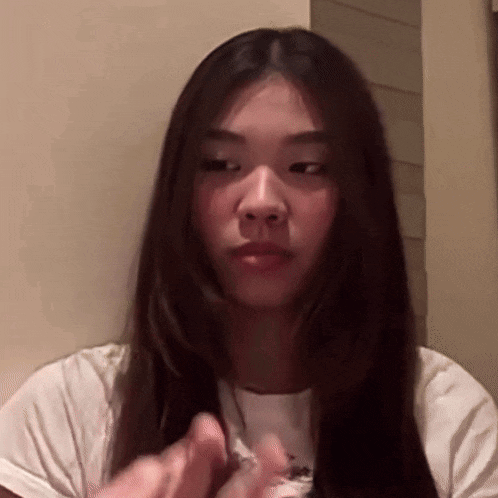 a woman with long dark hair is sitting in front of a wall and making a funny face .