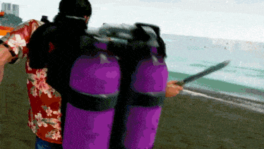 a man in a floral shirt is carrying purple scuba tanks on a beach