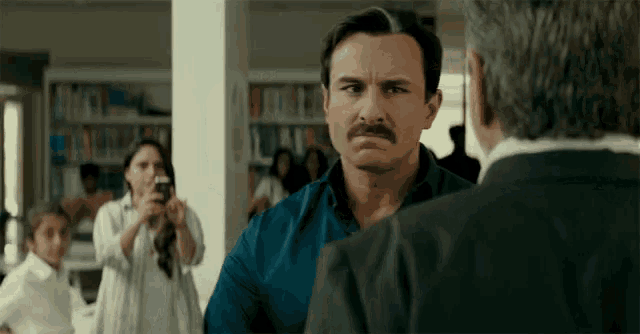 a man with a mustache is talking to another man in a library while a woman takes a picture of him .