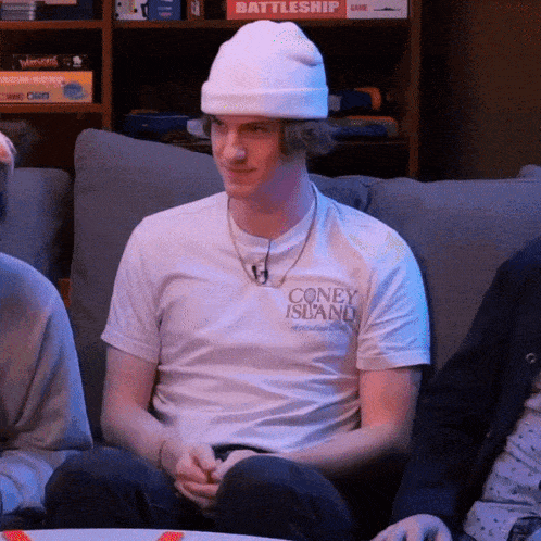 a man wearing a white beanie and a coney island shirt sits on a couch
