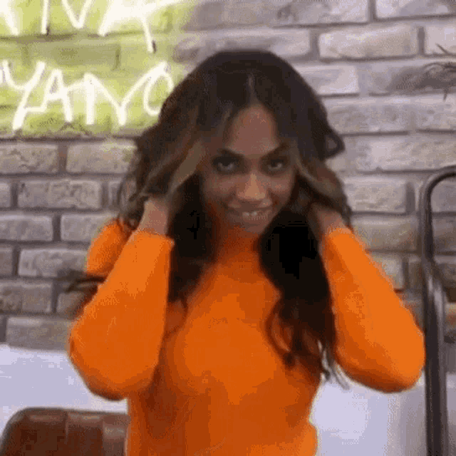 a woman in an orange sweater is standing in front of a brick wall holding her hair .