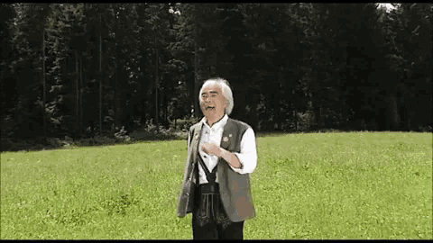 a man in a green vest stands in a grassy field with trees in the background