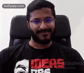 a man with a beard is sitting in a chair wearing glasses and a black t-shirt .