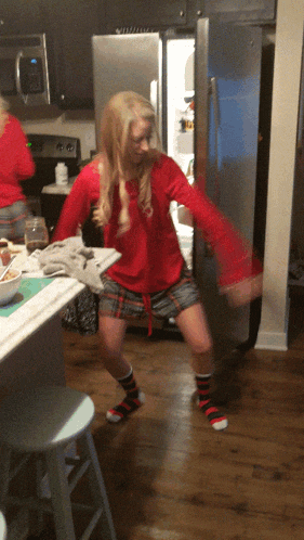 a woman in a red shirt and plaid shorts is dancing in front of a fridge