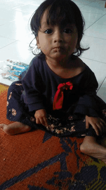 a little girl wearing a purple shirt and a red bow sits on a rug