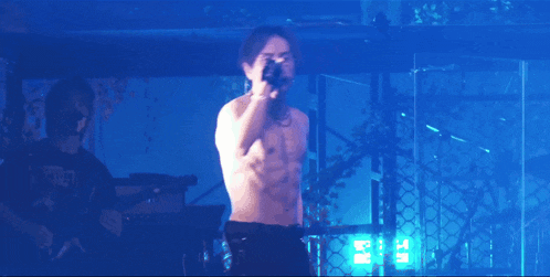 a shirtless man singing into a microphone on a stage with a blue background