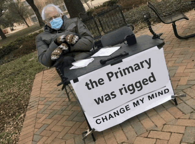 a man wearing mittens sits behind a sign that says the primary was rigged