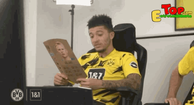 a man sitting in a chair reading a book with the number 187 on his shirt