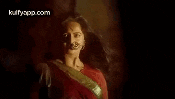 a woman in a red saree with her hair blowing in the wind is standing in a dark room .