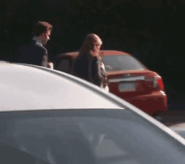 a man and a woman are walking past a red car .