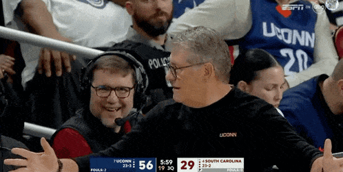 a man wearing a uconn shirt talks into a microphone while watching a game