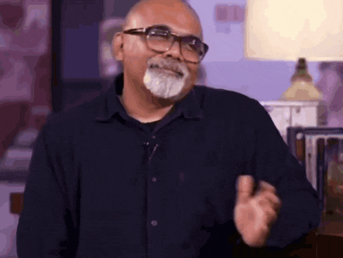 a man with glasses and a beard is wearing a black shirt and making a gesture .