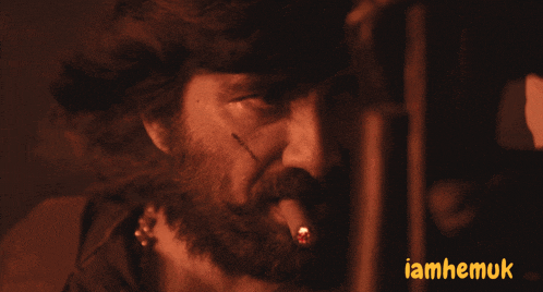 a man with a beard and a tattoo on his face is smoking a cigarette with the words iamhemuk behind him