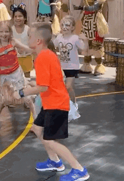 a boy in an orange shirt is dancing in front of a group of children in costumes .