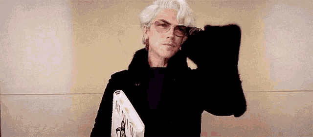 a man with white hair and glasses is standing in front of a wall holding a folder .