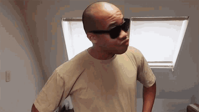 a bald man wearing sunglasses and a tan shirt is standing in front of a window
