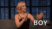 a woman in a black dress is sitting on a couch and making a gesture with her hands while saying boy .