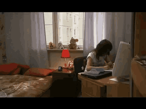a girl sits at a desk in front of a window with stuffed animals on the window sill