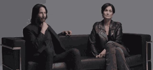 a man in a suit and a woman in a leather jacket are sitting on a couch .