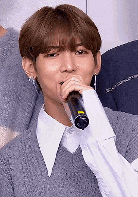 a young man is singing into a microphone while wearing a grey sweater and a white shirt .