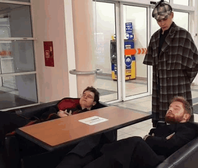 a man in a plaid coat is standing next to two men sleeping on a table