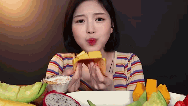a woman is eating a slice of mango while sitting at a table filled with fruit .