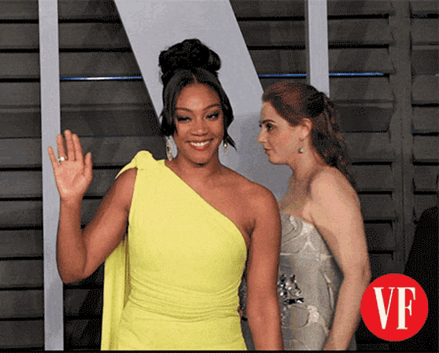 a woman in a yellow dress waves to another woman with a vf logo in the corner