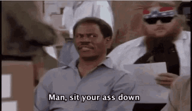 a man in a blue shirt is sitting in a crowd of people and says `` man , sit your ass down '' .