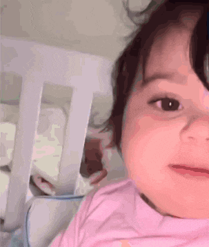 a baby girl in a pink shirt is laying in a crib looking at the camera .